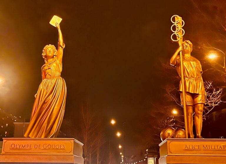 Statues illuminées d'Olympe de Gouges et d'Alice Milliat de nuit sur la rue de la Chapelle.